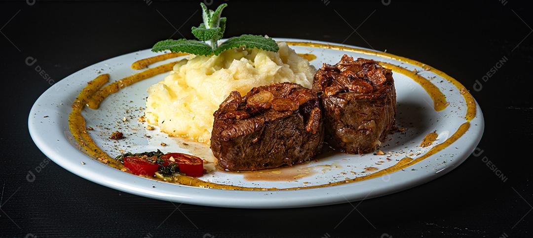 Filé Mignon Com Cebola Frita e Purê De Batata, Decorado Com Tomate Cereja e Molho De Mostarda Em Fundo Preto.