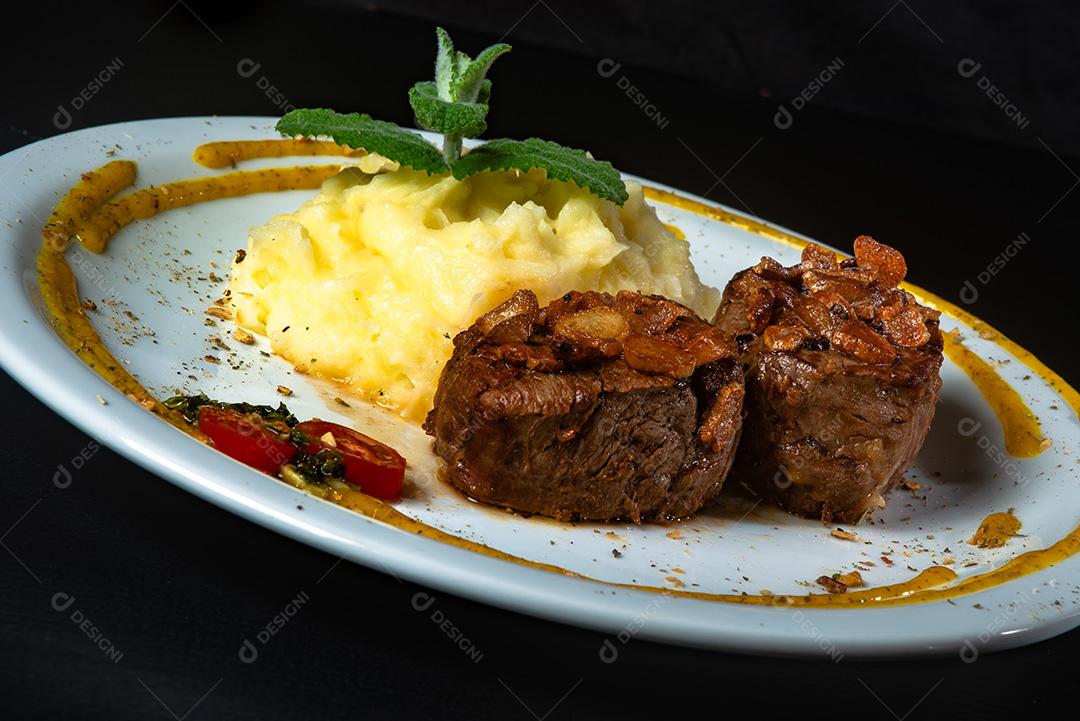 Filé Mignon Com Cebola Frita e Purê De Batata, Decorado Com Tomate Cereja e Molho De Mostarda Em Fundo Preto.