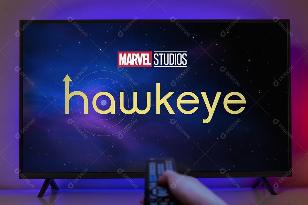 Logo Hawkeye na Tela da Tv e Mão Segurando Um Controle Remoto Imagem JPG