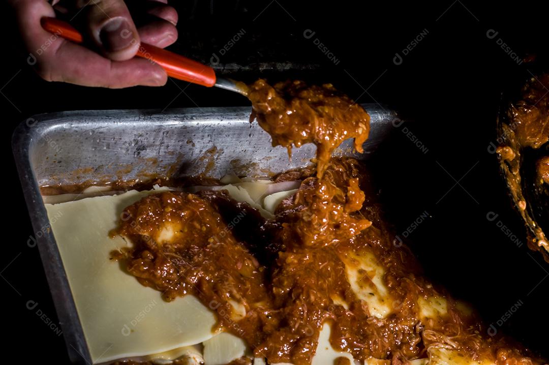 lasanha sendo montada à mão colocando molho e fundo escuro Imagem JPG