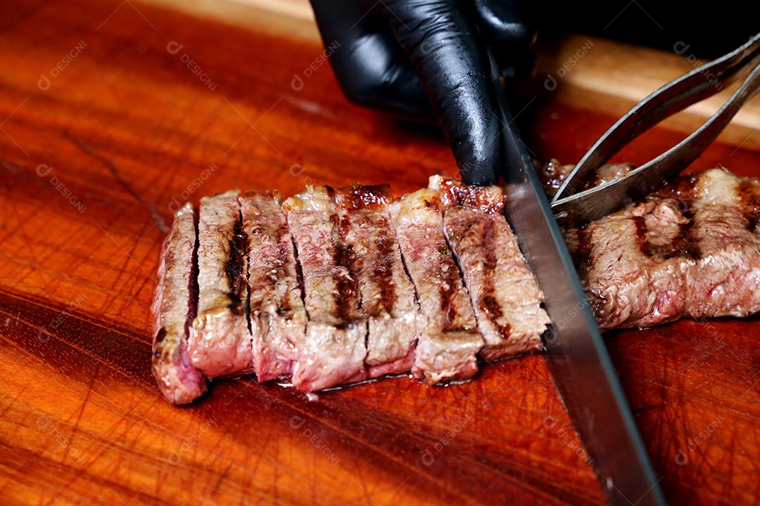 Picanha ou Bife de Alcatra. Mão com luvas pretas cortando um bife suculento em uma placa de madeira JPG