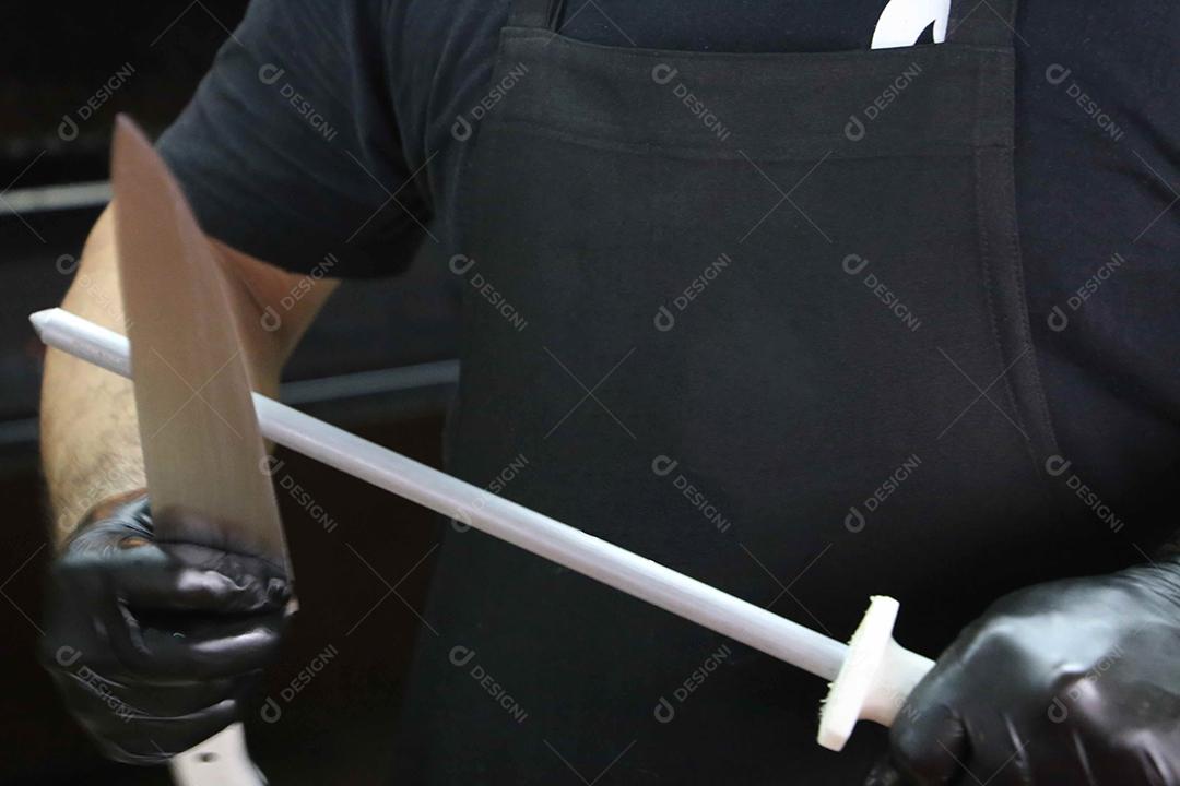 homem afiando uma faca de churrasco na cadeira. Com avental preto ao fundo JPG