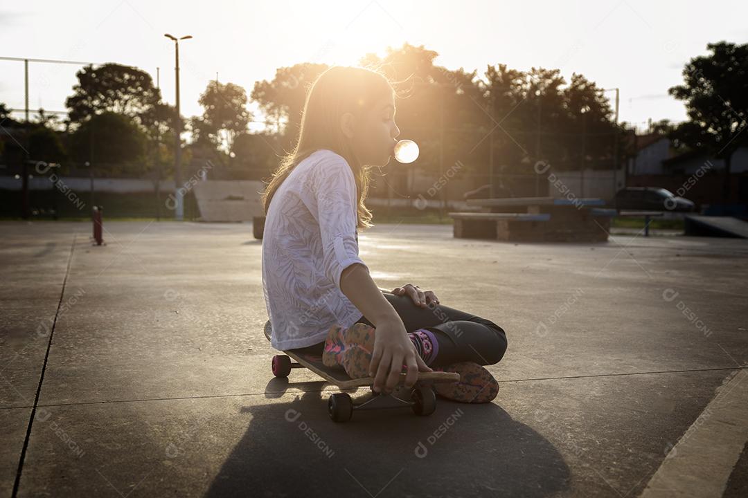 Linda loira sentada em cima do skate no parque ao pôr do sol Imagem JPG
