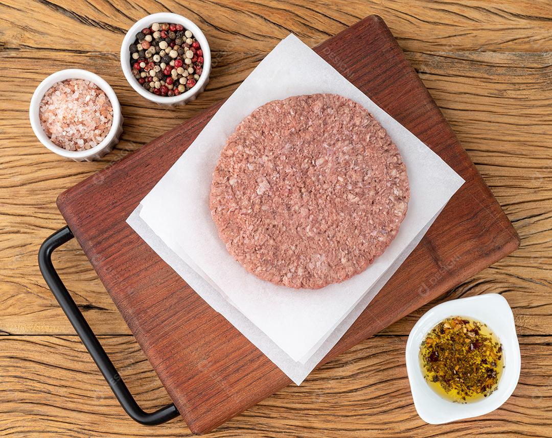 Hambúrguer de carne crua sobre tábua de madeira com temperos.