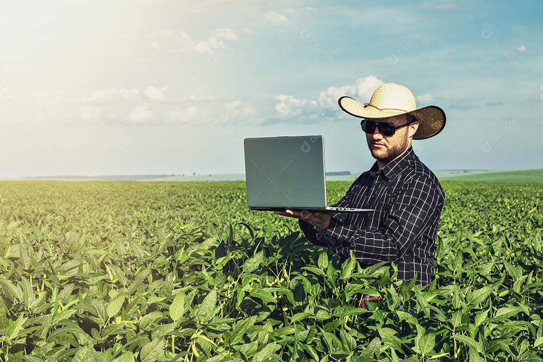 Jovem agrônomo de chapéu segurando o notebook no campo de soja.Imagem JPG