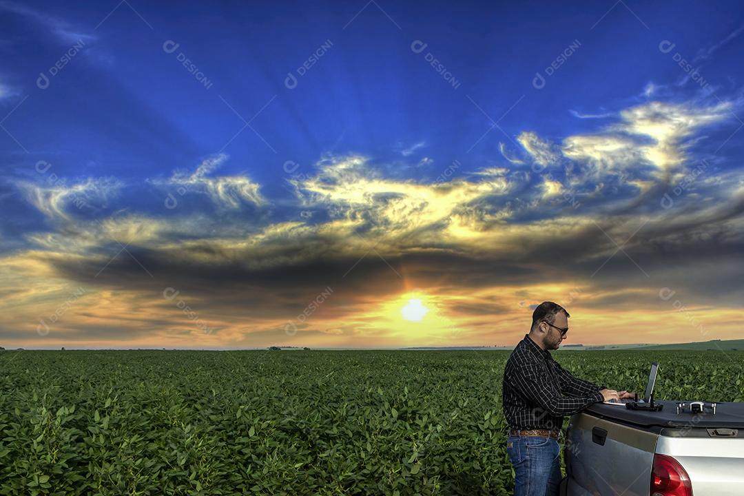 Agricultor de pé no campo de soja olhando para notebook e drone Imagem JPG