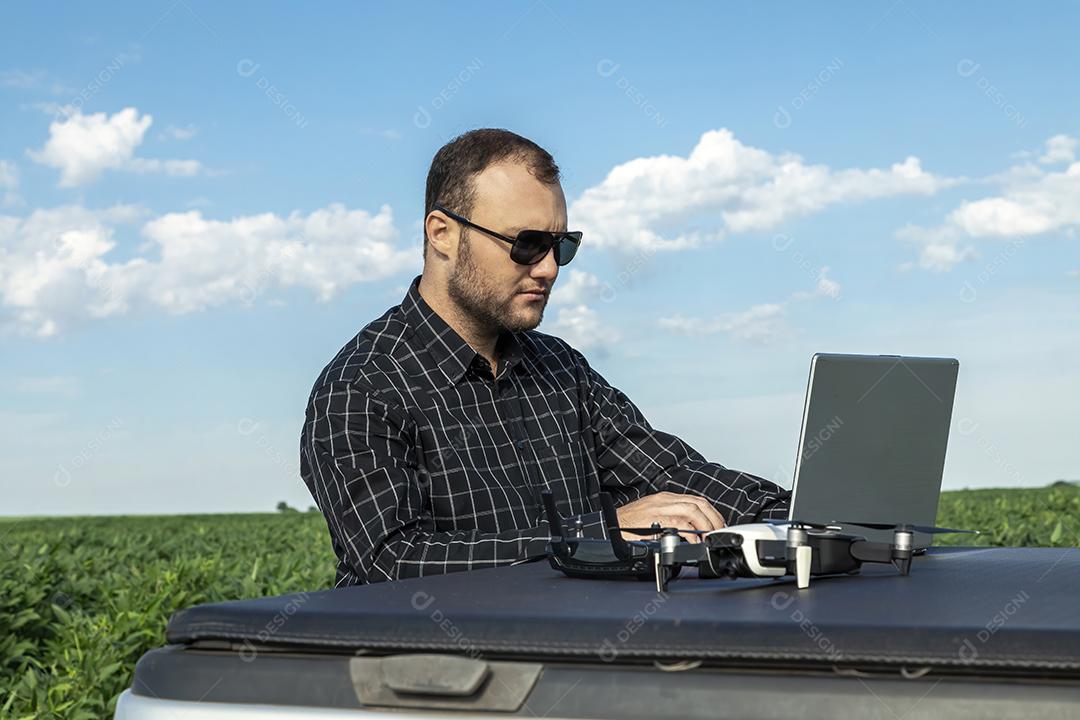 Agricultor de pé no campo de soja olhando para notebook e drone Imagem JPG