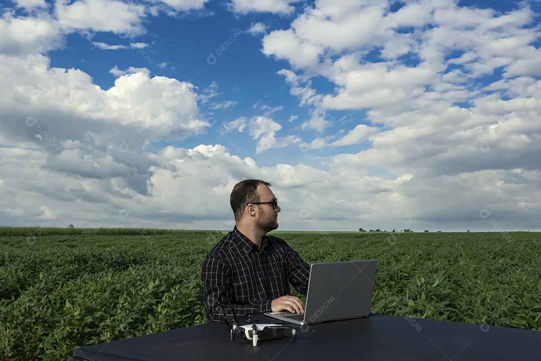 Agricultor de pé no campo de soja olhando para notebook e drone Imagem JPG