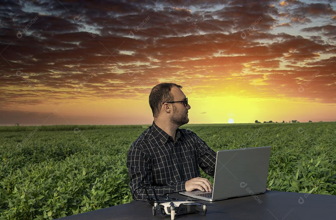 Agricultor de pé no campo de soja olhando para notebook e drone Imagem JPG