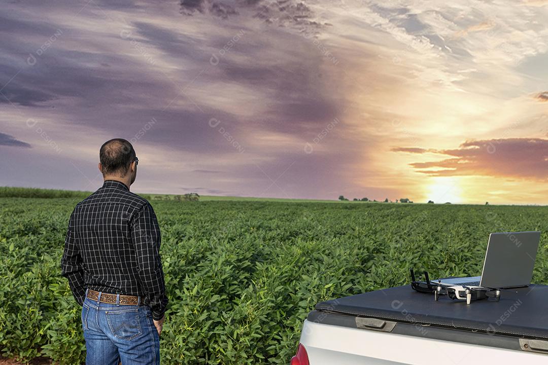Agricultor de pé no campo de soja olhando para notebook e drone Imagem JPG