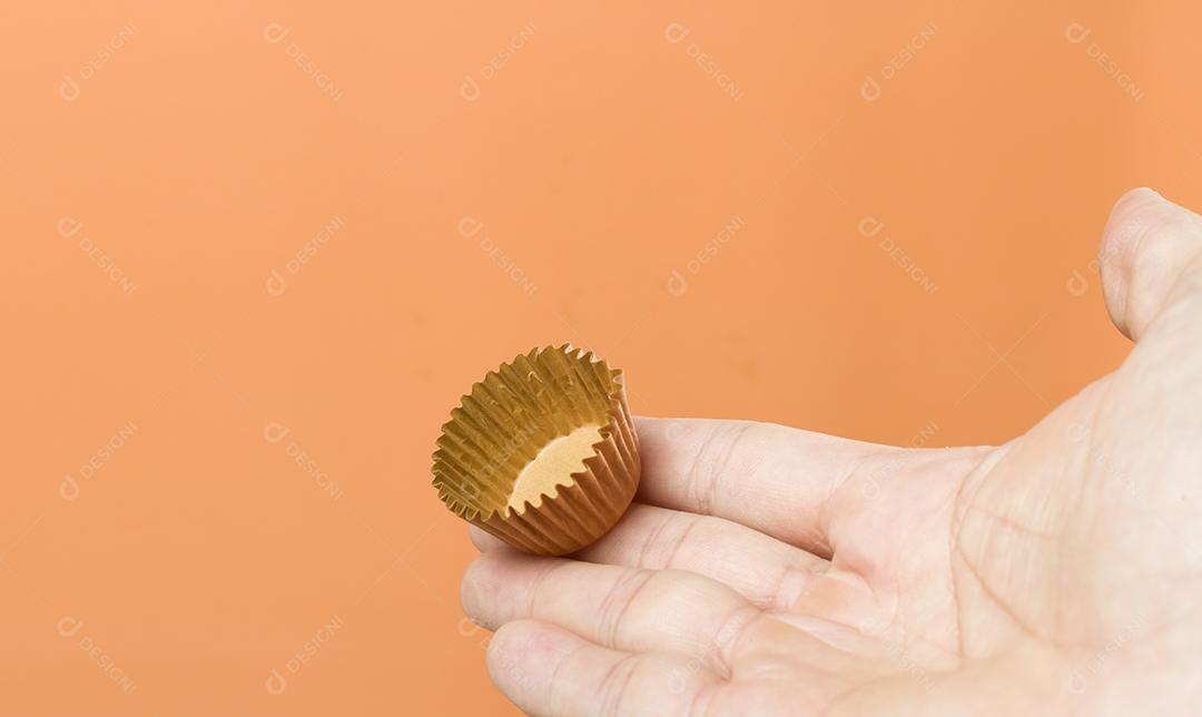 Mão de mulher segurando um brigadeiro vegano. Típico doce brasileiro. Fundo laranja.