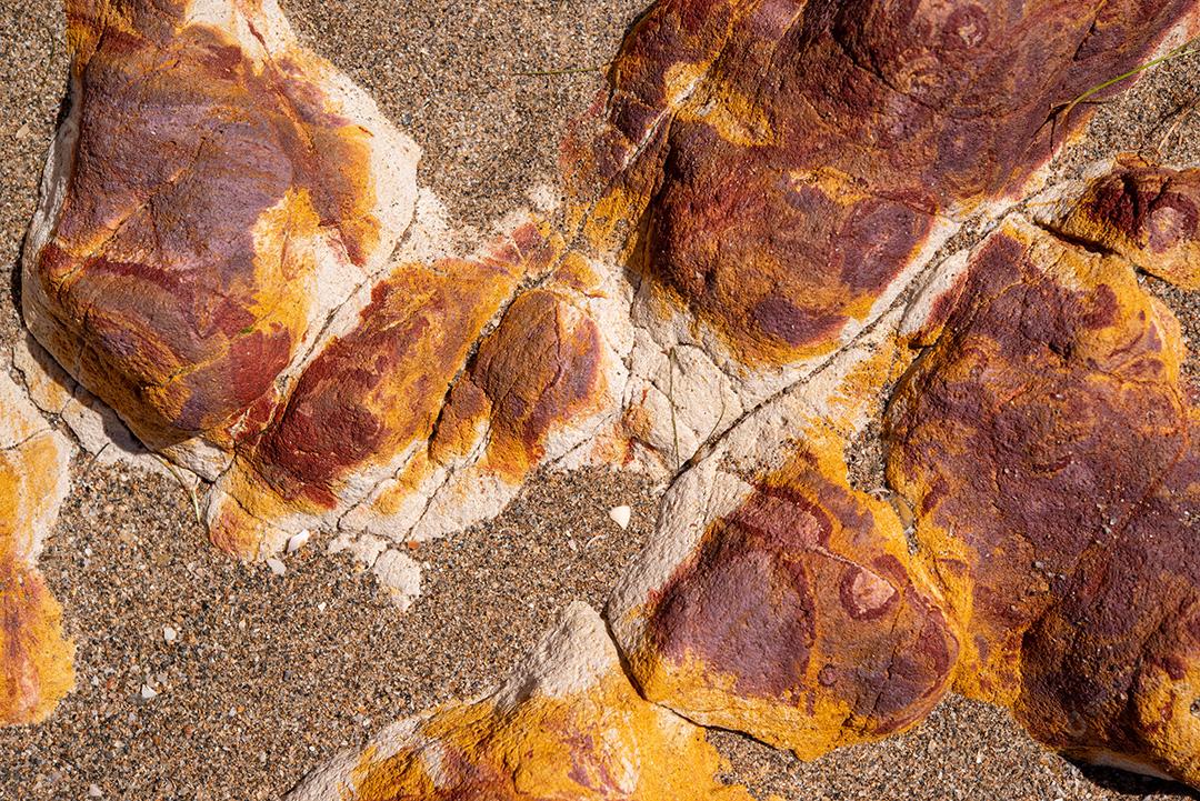 Areia colorida e rochas sedimentares, criando padrões abstratos na praia