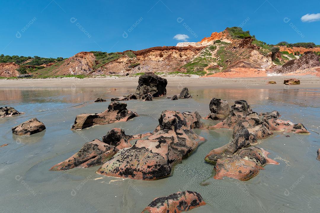 Pedras coloridas e areia na praia, ao fundo falésias com vegetação natural na Praia de Peroba