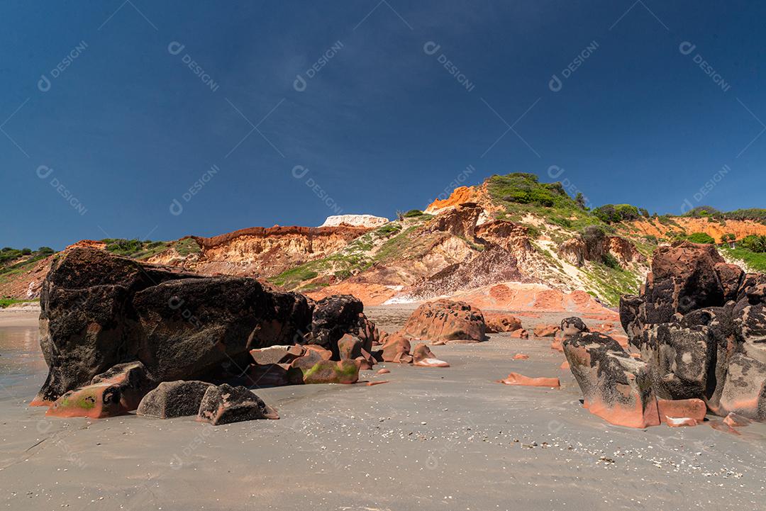 Pedras coloridas e areia na praia, ao fundo falésias com vegetação natural na Praia de Peroba