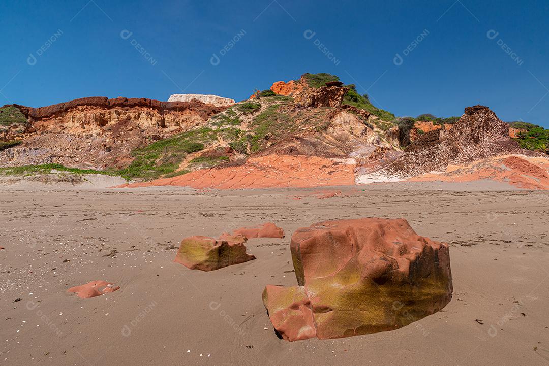 Pedras coloridas e areia na praia, ao fundo falésias com vegetação natural na Praia de Peroba
