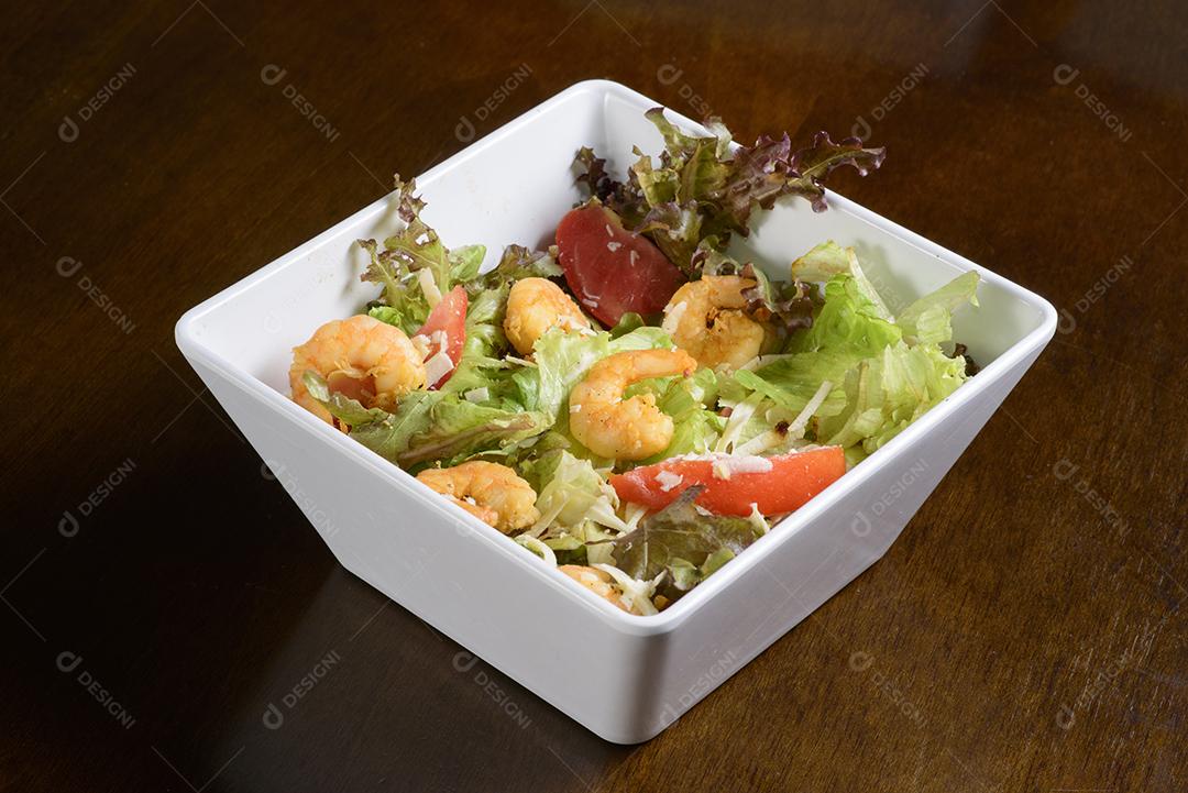 Salada verde com camarão em tigela branca na mesa de madeira.