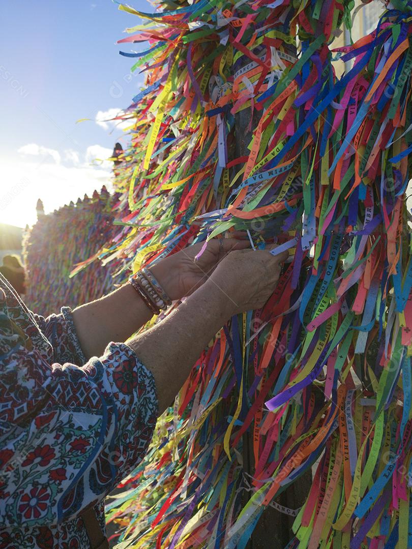 Mãos femininas amarrando fitas coloridas na grade da igreja do Bonfim