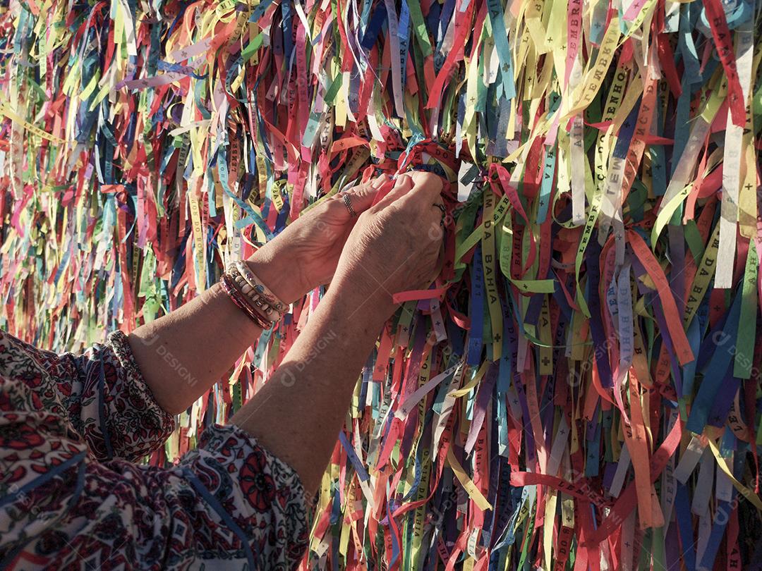 Mãos femininas amarrando fitas coloridas na grade da igreja do Bonfim