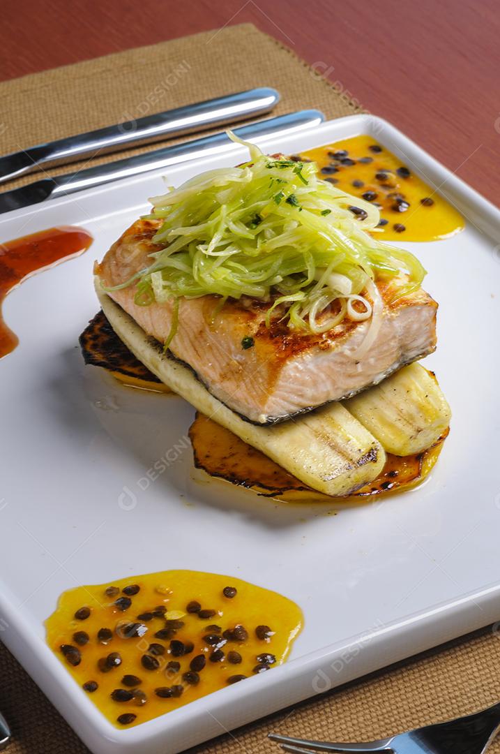 Filé de salmão com bananas e batatas em um prato quadrado branco servido à mesa.