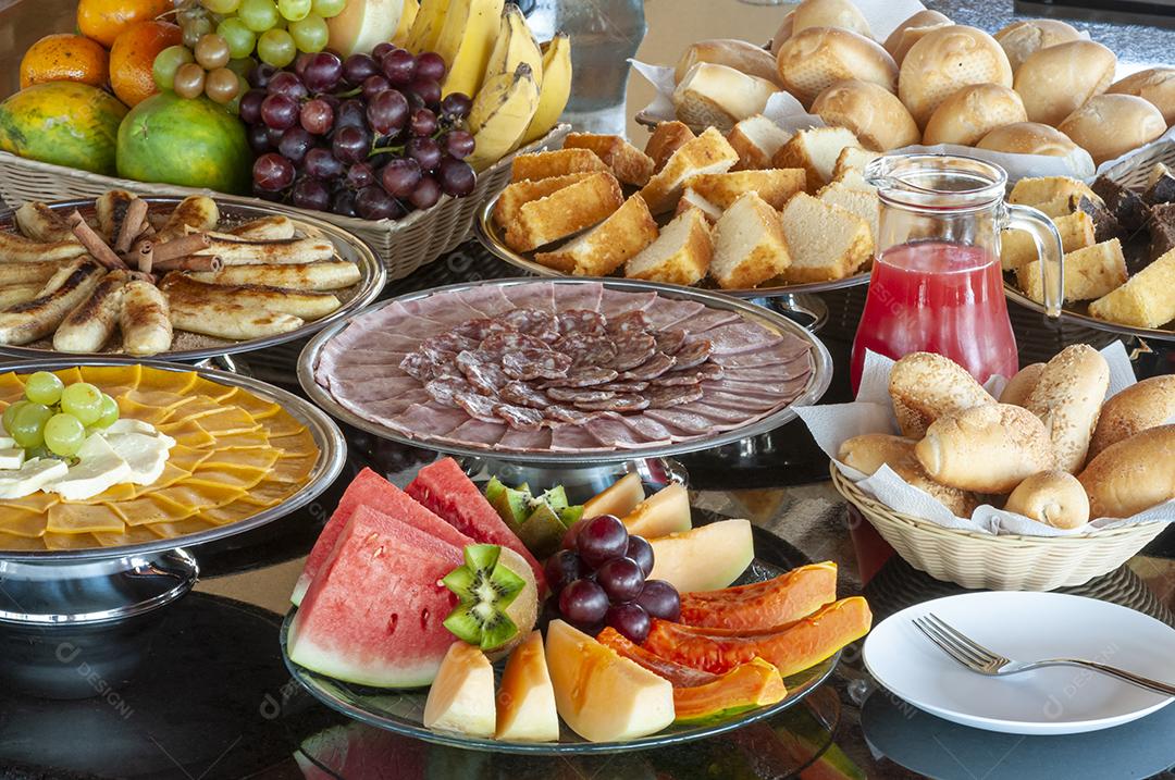 Mesa de café da manhã com salame, presunto, pão, frutas, queijo, bolo e suco.