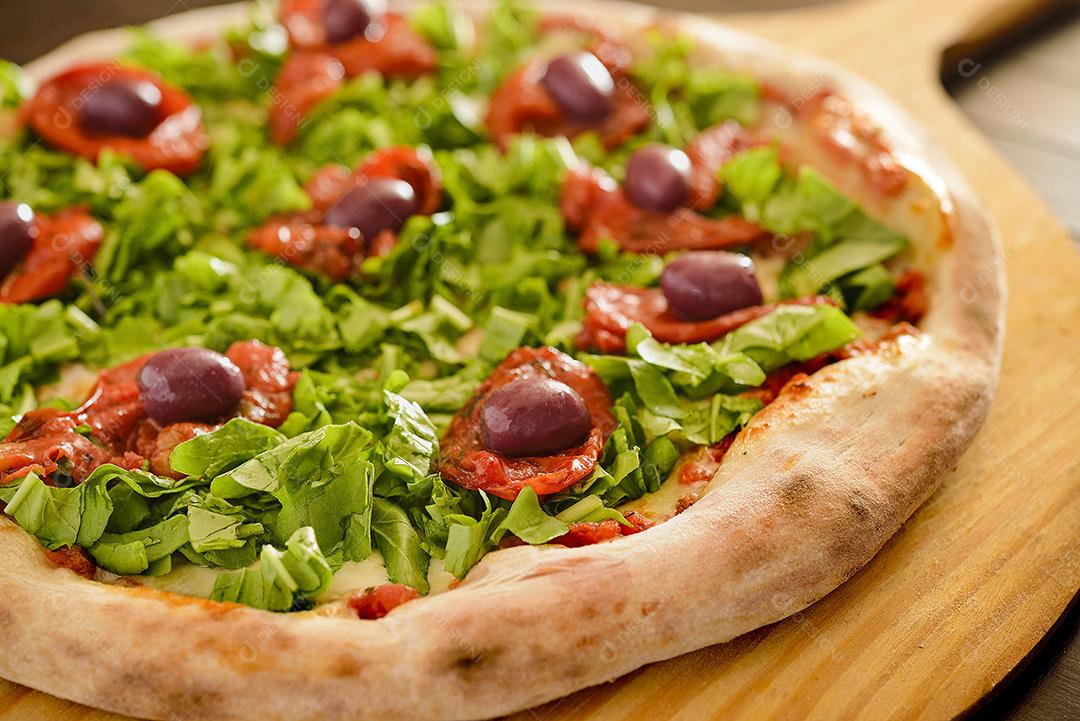 Pizza de rúcula com tomate seco e azeitonas em close-up de pá de madeira.