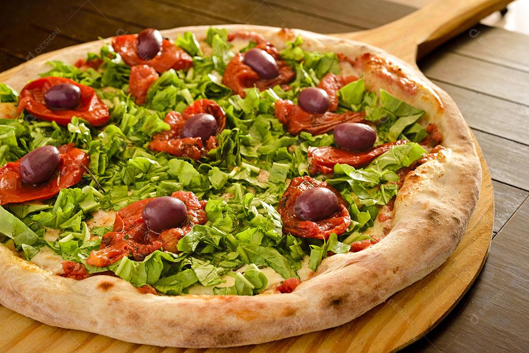 Arugula pizza with dried tomatoes and olives on wooden shovel close-up.