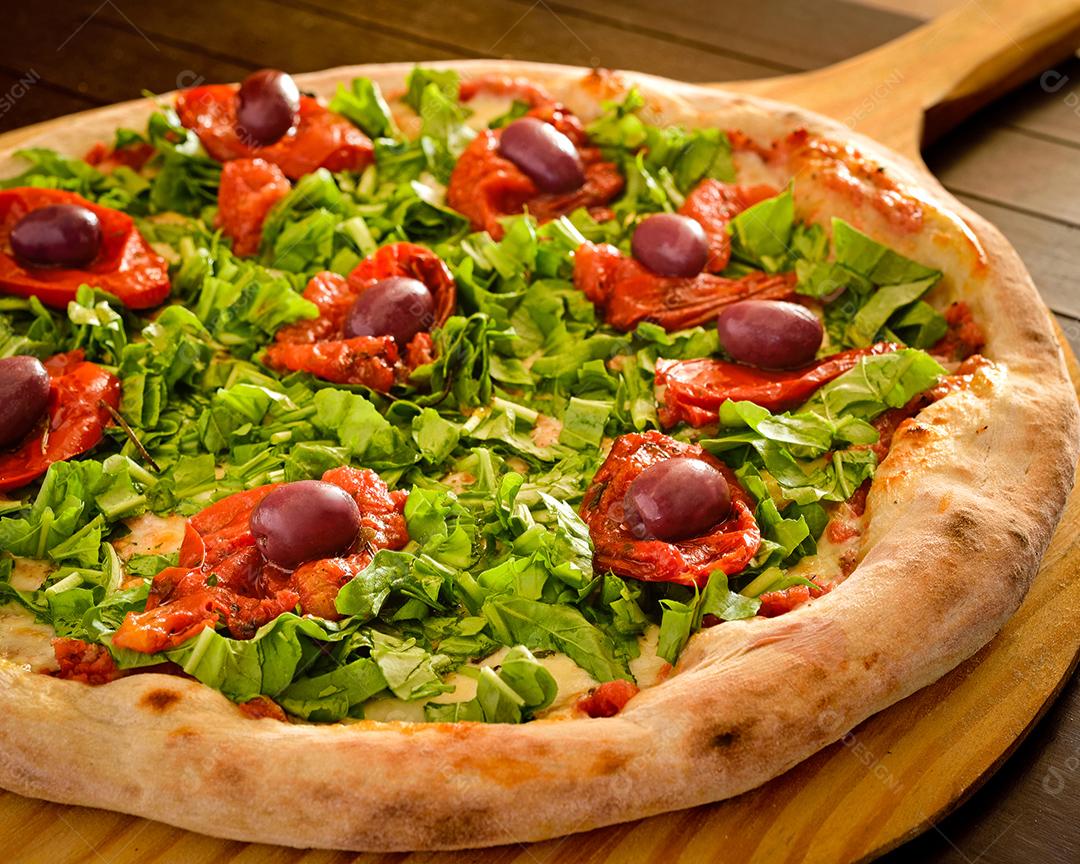 Pizza de rúcula com tomate seco e azeitonas em close-up de pá de madeira.