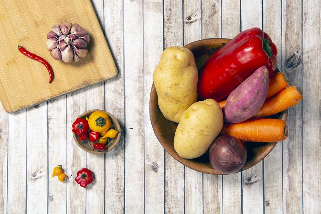 Batatas cruas, cenouras e cebolas em uma tigela ao lado de alho e pimentão em uma placa de madeira sobre a mesa de madeira.
