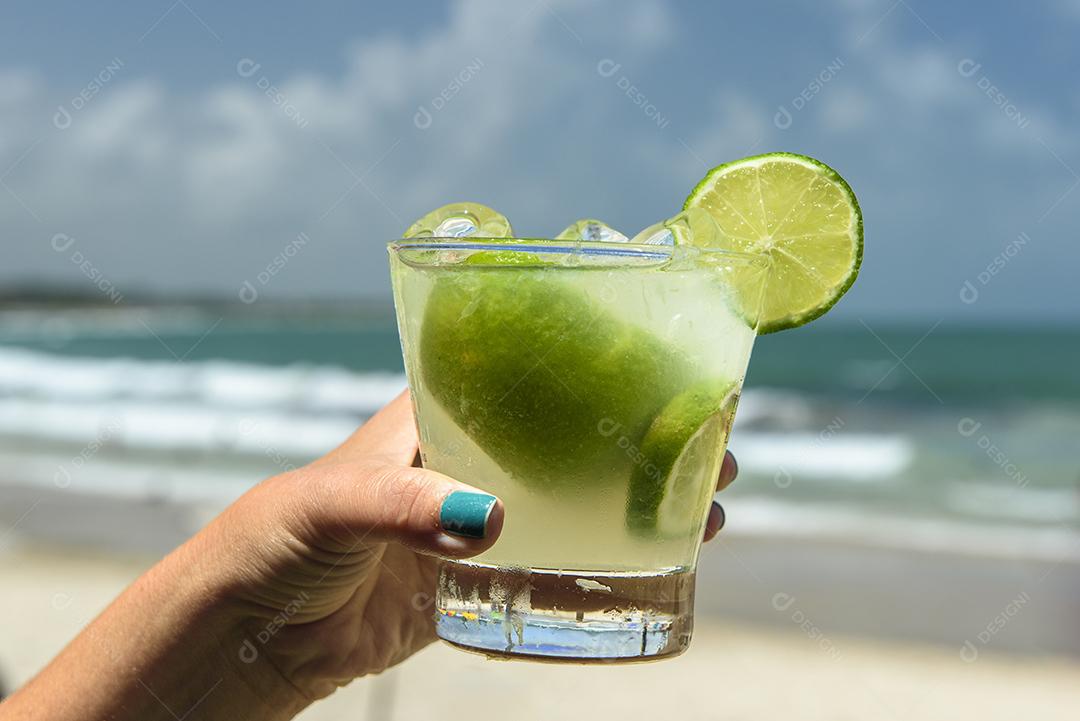 Beba caipirinha. Mão segurando uma bebida caipirinha em uma praia