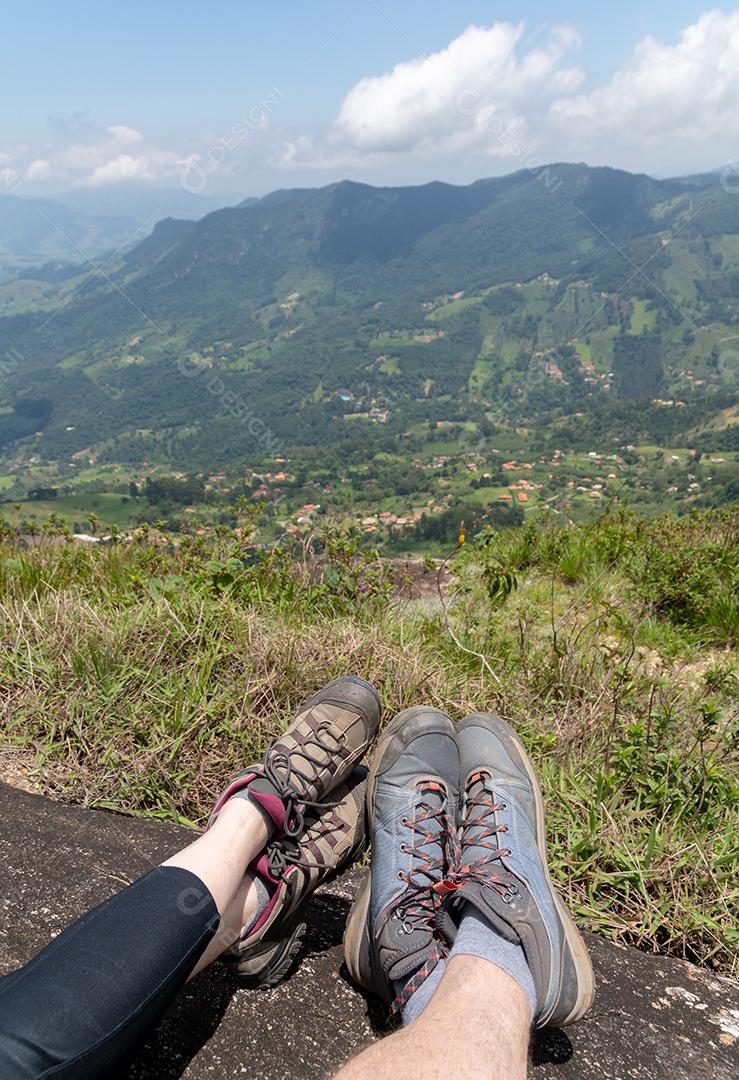 Casal descansando no topo de uma montanha mostrando os pés em frente a uma vista natural.