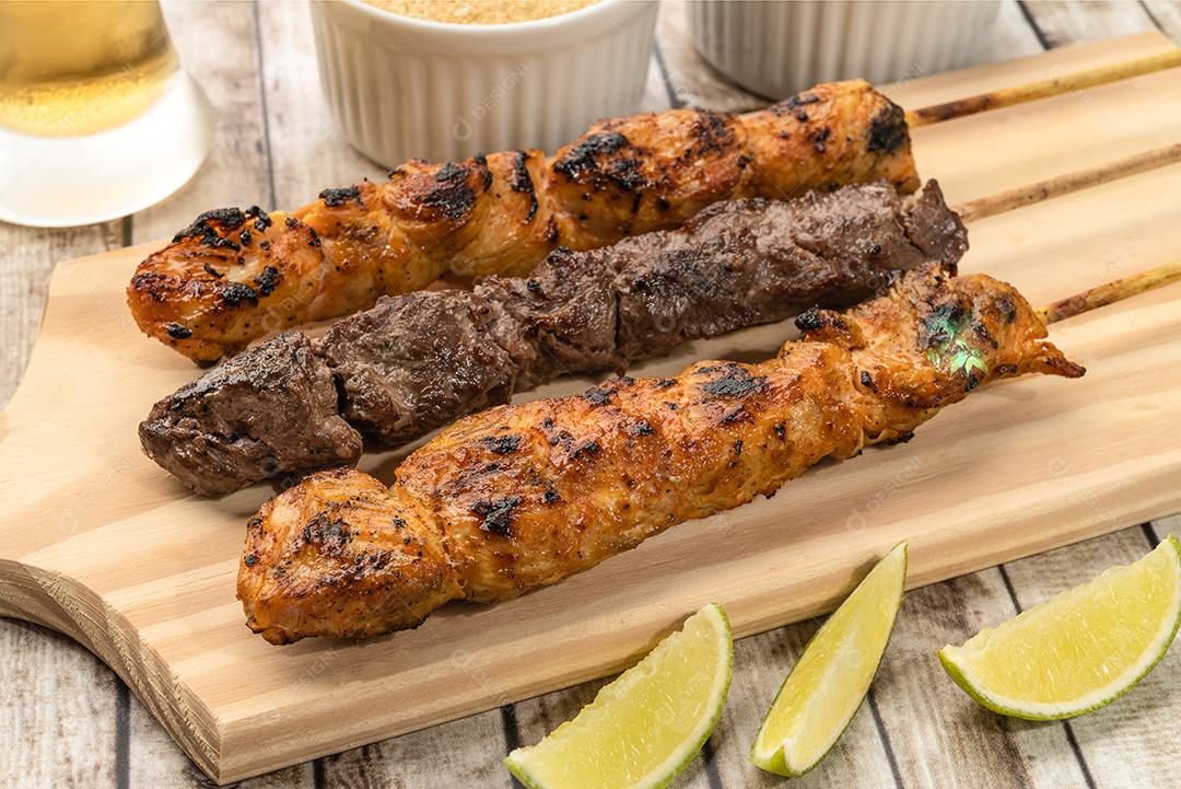 Espetos de frango e carne sobre tábua de madeira com farofa, limão