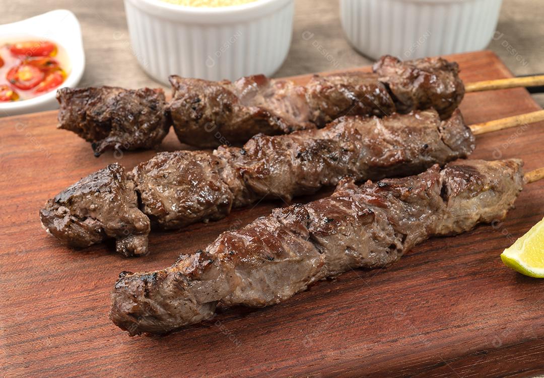 Espetos de carne sobre tábua de madeira com farofa, limão e pimenta