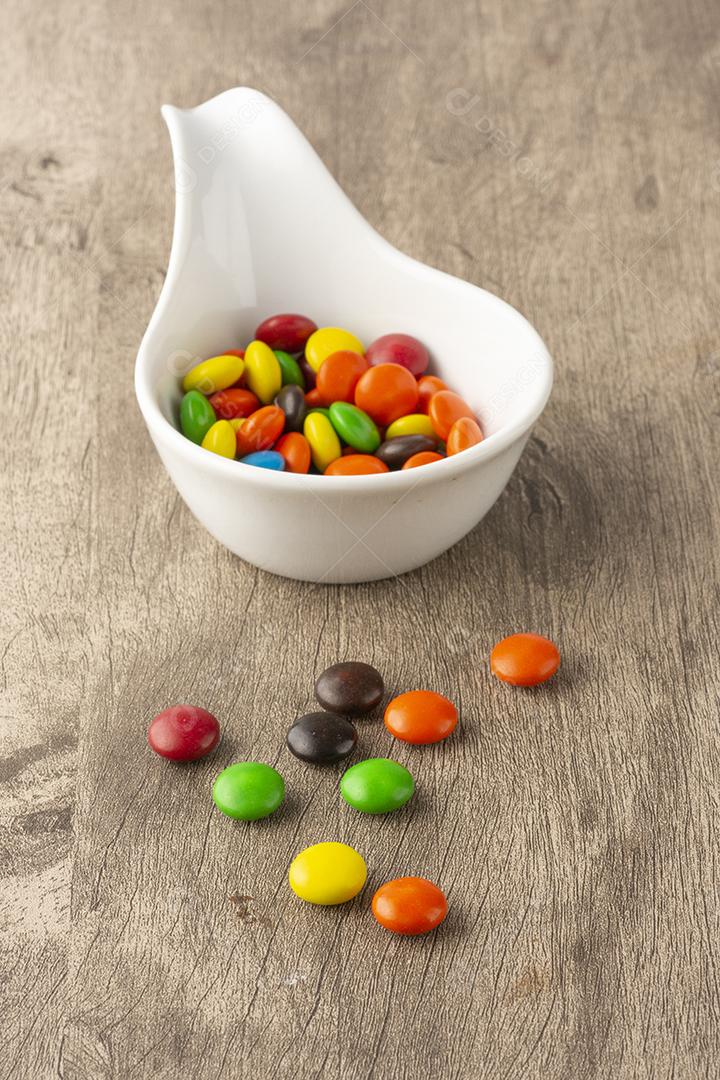 Doces coloridos em uma colher branca e espalhados sobre mesa de madeira