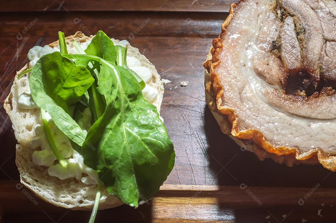 Porchetta, sanduíche de porchetta sendo montado em uma mesa de madeira, ao lado de folhas de pão e rúcula.