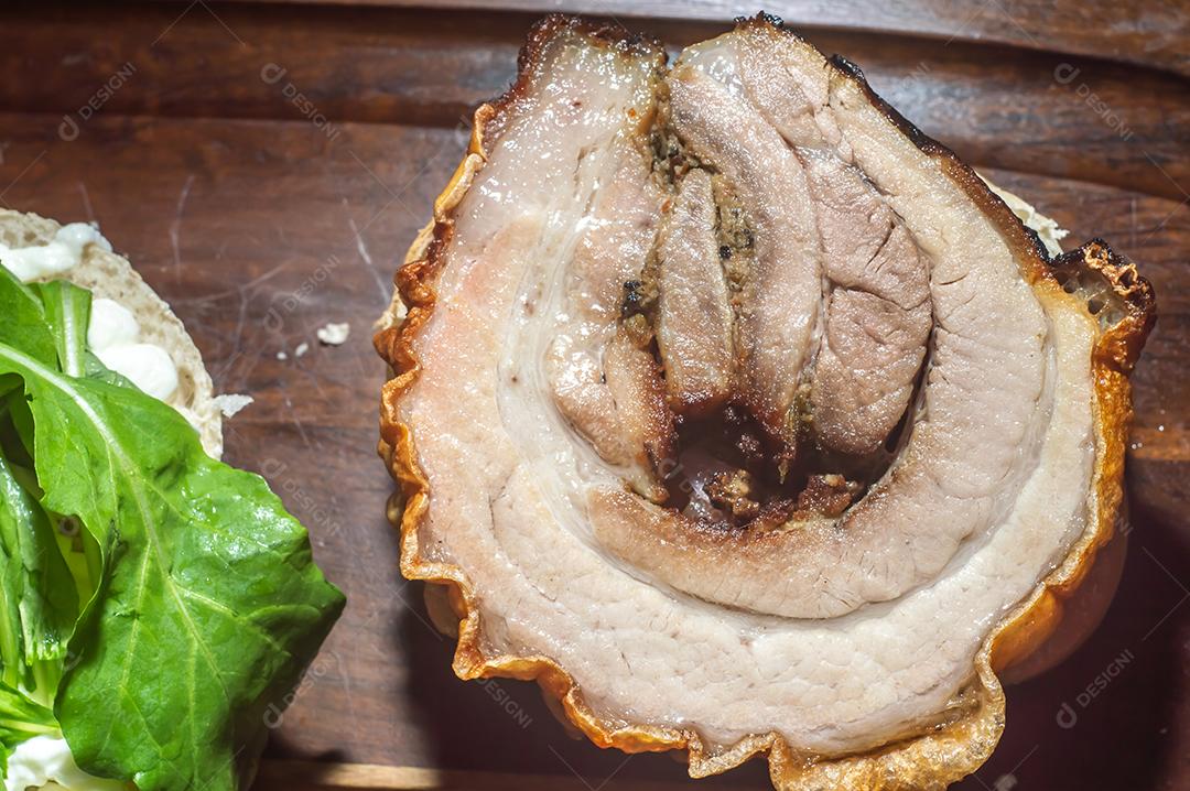 Porchetta, sanduíche de porchetta sendo montado em uma mesa de madeira, ao lado de folhas de pão e rúcula.