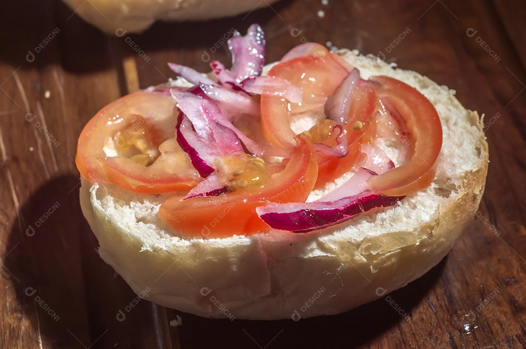 Pão com tomate e cebola fundo de um lanche sendo montado em uma mesa de madeira.