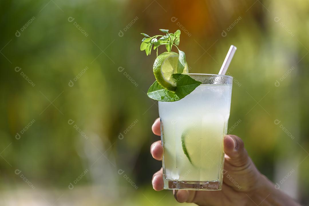 Brasileira bebe caipirinha. Mão segurando uma bebida de caipirinha com folhagens borradas ao fundo