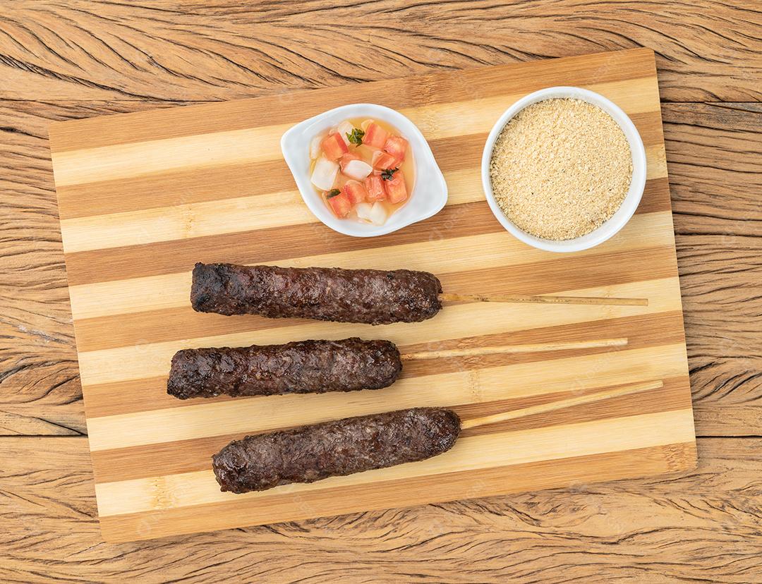 Espetinhos de Kafta de carne sobre tábua de madeira com farofa e vinagrete