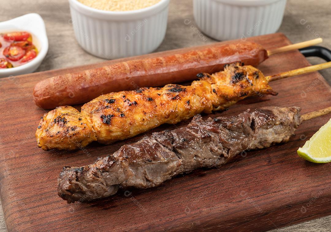 Espetos de carne, frango e salsicha de porco sobre tábua de madeira