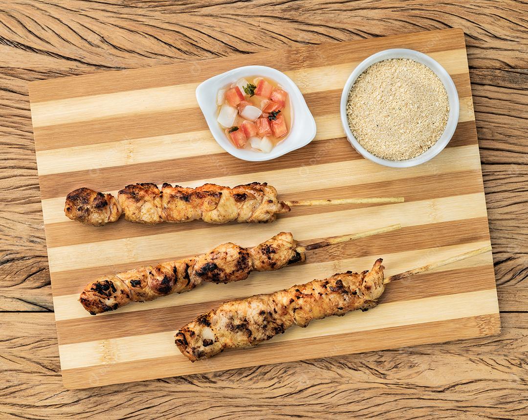 Espetos de frango sobre tábua de madeira com farofa e vinagrete