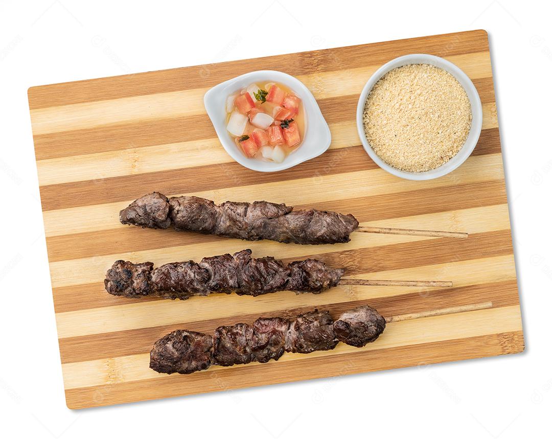 Espetos de carne sobre tábua de madeira com farofa e vinagrete