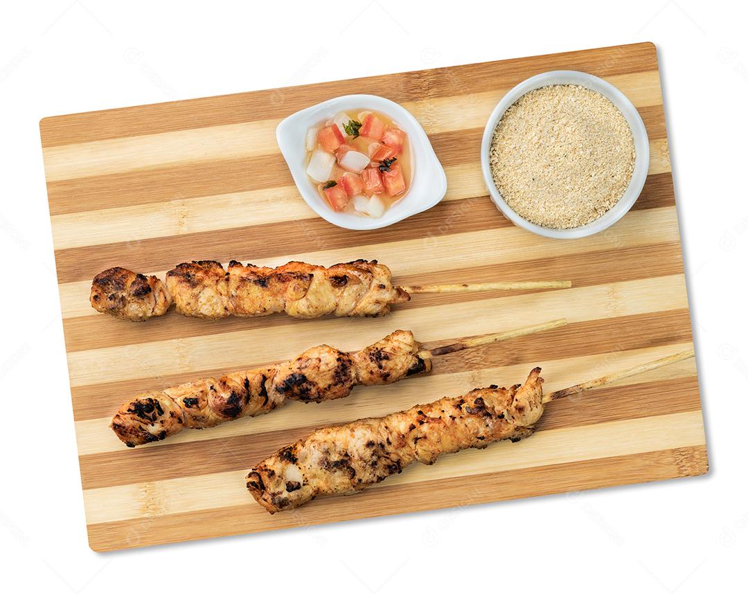 Espetos de frango sobre tábua de madeira com farofa e vinagrete