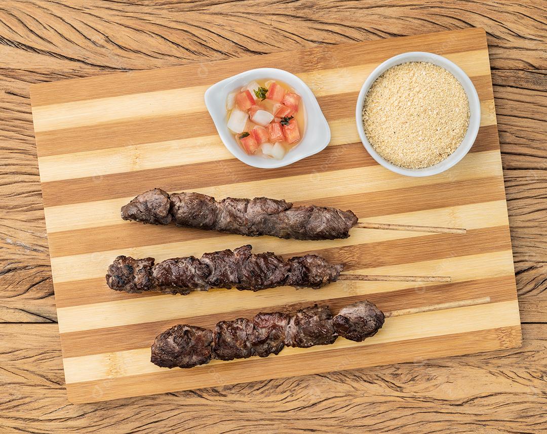 Espetos de carne sobre tábua de madeira com farofa e vinagrete
