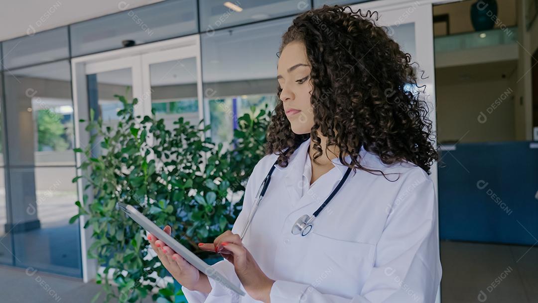 Jovem médica latina usa uniforme branco, usando tablet digital