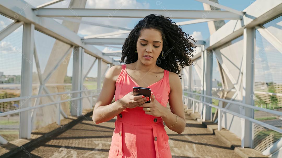 Jovem mulher latina de cabelo encaracolado andando usando o celular. Mensagens de texto