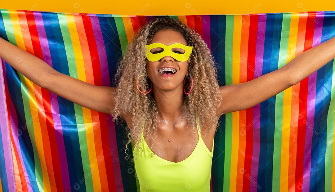 Mulher jovem de cabelo encaracolado fantasiada curtindo a festa de carnaval cobrindo com a bandeira do orgulho lgbt.