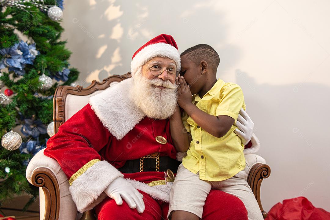 Um menino africano sussurrando no ouvido do Papai Noel. Contando um segredo