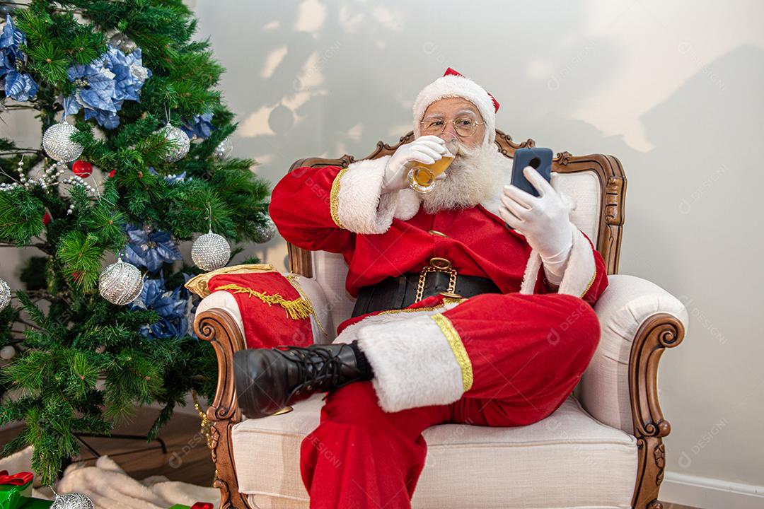 Papai Noel bebendo um copo de cerveja enquanto usava o celular.