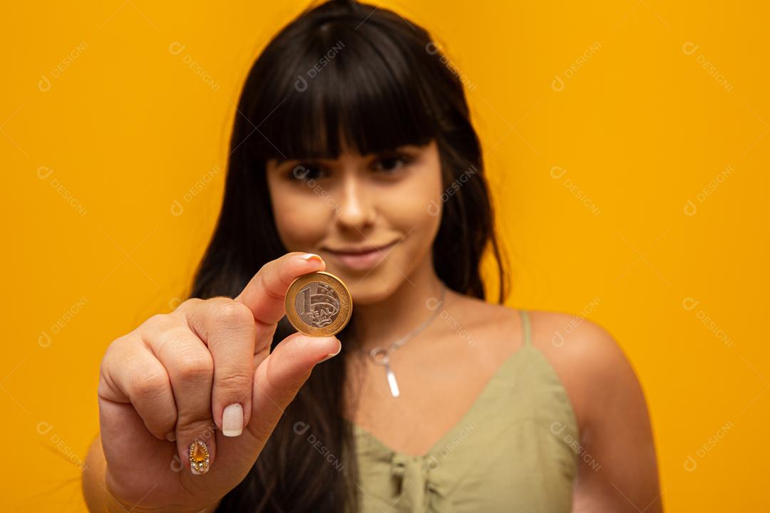 Mão de mulher jovem segurando uma moeda real do Brasil em fundo amarelo. Conceito de finanças.