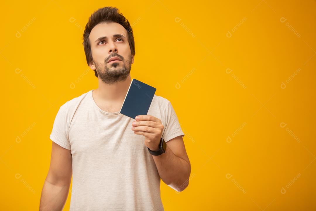 Jovem segurando um passaporte, cartão de trabalho ou documento, pensando. Em fundo amarelo